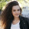 Portrait of beautiful young woman with clean healthy skin and long curly dark hair wearing trendy leather jacket relaxing outdoors on sunny day. People, leisure and modern urban lifestyle concept